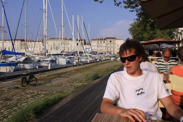 A man in white t-shirt sitting at wooden picnic table by water, boats docked on other side of canal. 