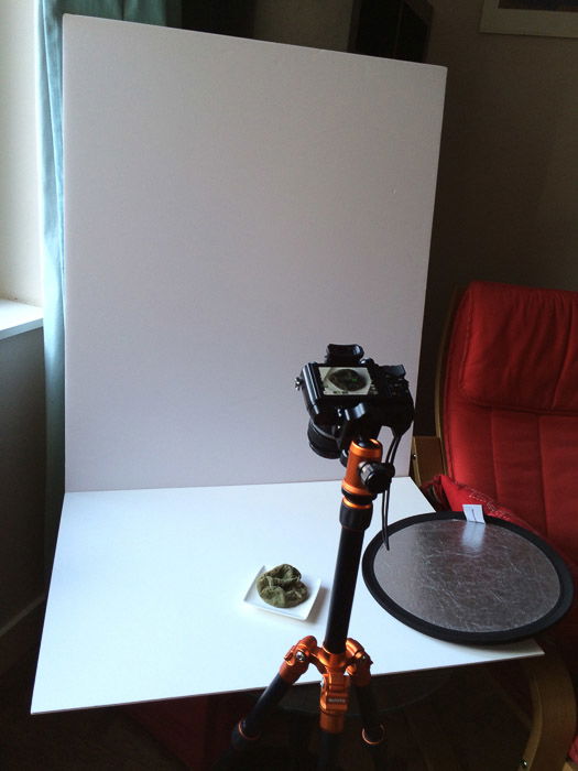 View of a high key set up for still life photography with white cardboard background tabletop, camera, tripod, and reflector