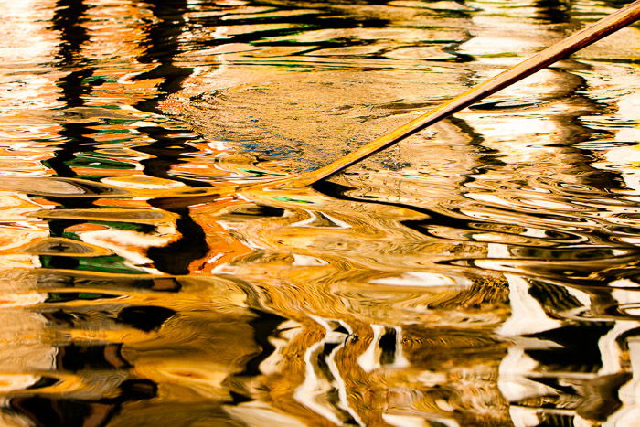 An oar breaking the surface of a river casting a choppy reflection. Night street photography in color.