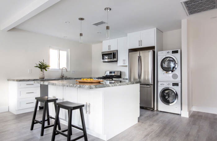 picture of a kitchen, showing the island in the middle with chairs, a double-door fridge and washer/dryer ensemble in the back