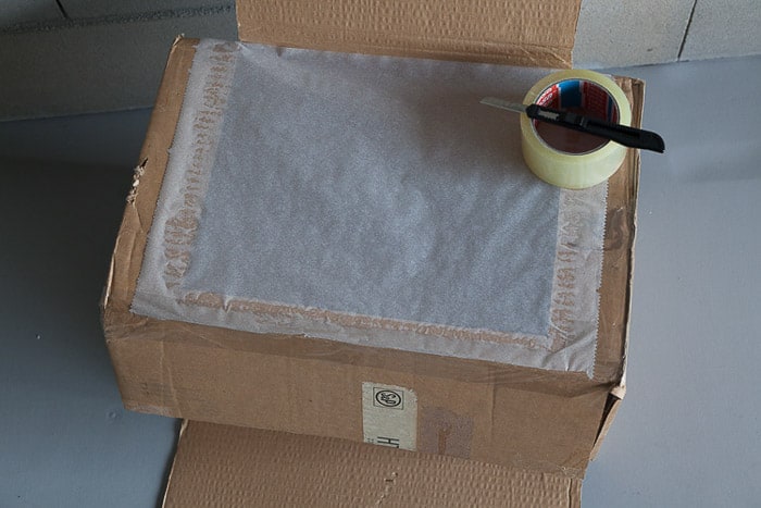 Overhead shot of a cardboard box with wax paper cover, tape, and X-Acto knife you need to make a DIY light box