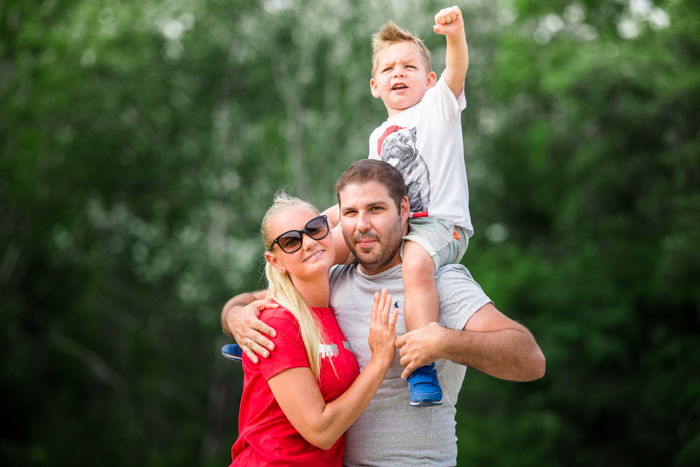 Parents standing in an embrace outside with their young toddler sitting on the father's shoulders with one arm raised and fist clenched for a family photo