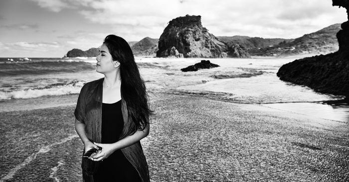 Black and white travel photo of a girl walking on Pina beach, New Zealand, rough waves and rocky coastline behind her