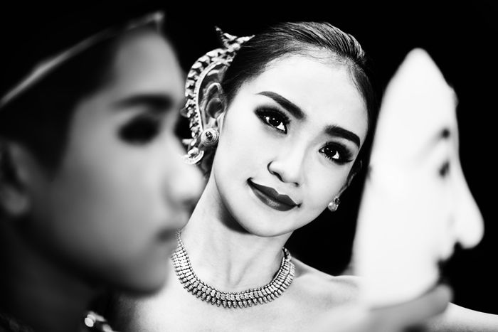 Black and white portrait of a girl in traditional costume smiling towards the camera, framed by a blurred figure in the foreground holding a mask, black and white photography