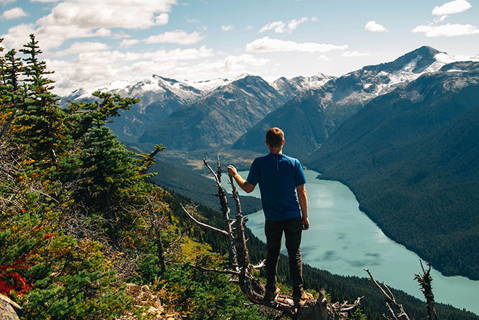 A man is blue t-shirt standing in the foreground of a mountainous landscape with his back turned to the camera