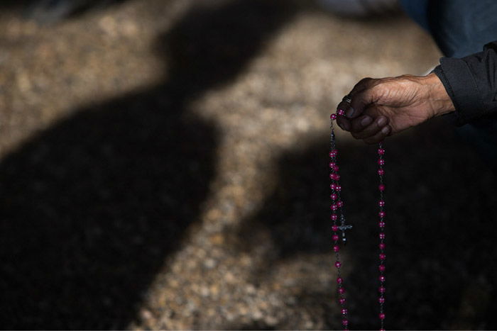 a hand holding rosary beads against a shadowing background. Photo by Amber Bracken.