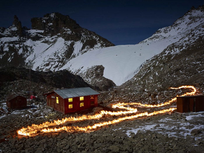 Photo of a firetrail by a woodenhouse in a snowy mountainous landscape by Simon Norfolk. 