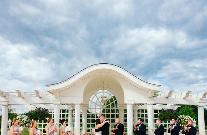 Photo of an outdoor bridal party showing bridesmaids and groomsmen applauding as bride and groom kiss. 