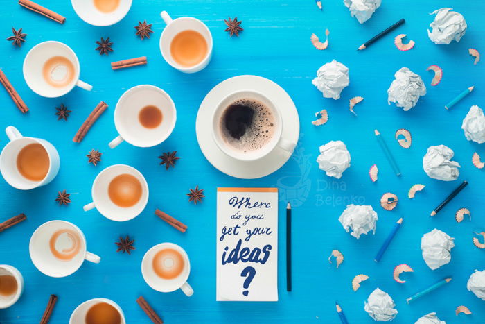 Overhead still life of coffee cups, cinnamon sticks, balls of paper and pencils with a notepad with the words 'where do you get your ideas' on sky blue background