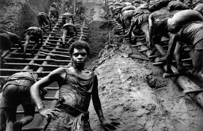 Man standing facing the camera at the bottom of a wooden ladder as other men climb up