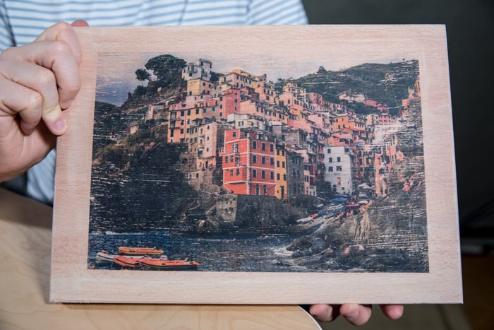 A man holding a wooden board with a colorful coastal town photo transferred to it