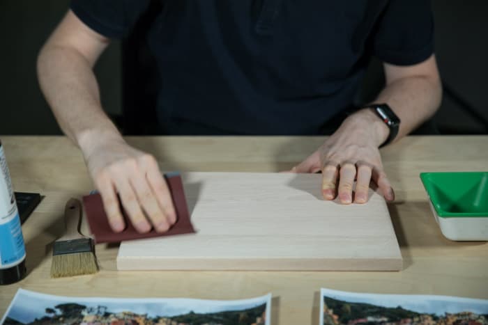 A man sits at a wooden table, sanding a wooden board to prepare the surface for to transfer photo to wood