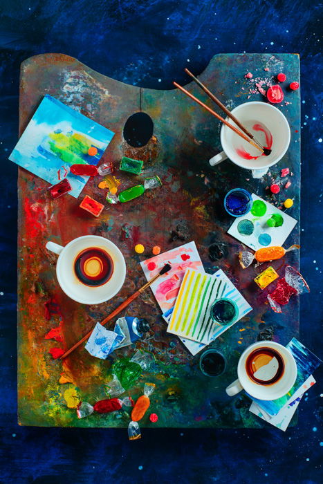 A bright artistic themed still life flat lay with a painters palette covered with paint, cups, paintbrushes and other art materials