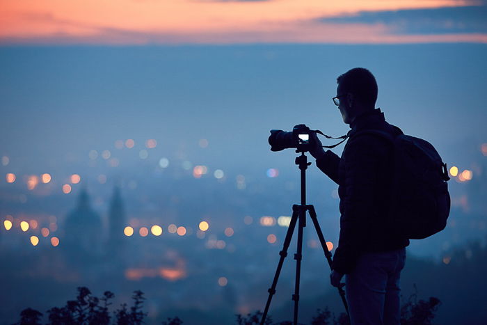 silhouette of photographer on a hill with camera on tripod taking a photo of a city at dusk