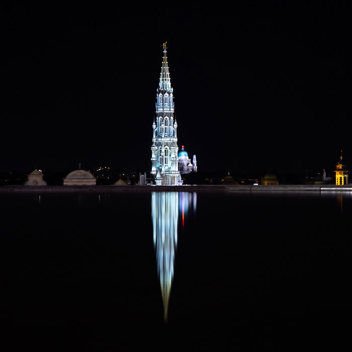 A reflection on the fountain in Mons des Arts against a black night sky. Town hall, Grand Palace, and Basilica of the Sacred Heart in Brussels in the background.