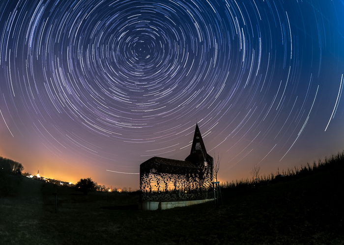 silhouette of the See Through Church lit warmly from behind, beneath a star trail and an orange blue dusk