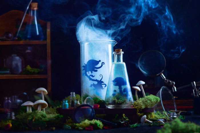 Mystical still life featuring glass bottles with silhouettes of tiny cutout characters inside and smoke billowing out