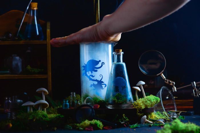 Placing a hand over a jar to trap smoke for a still life with glass bottles and silhouettes of tiny cutout characters inside and smoke billowing out