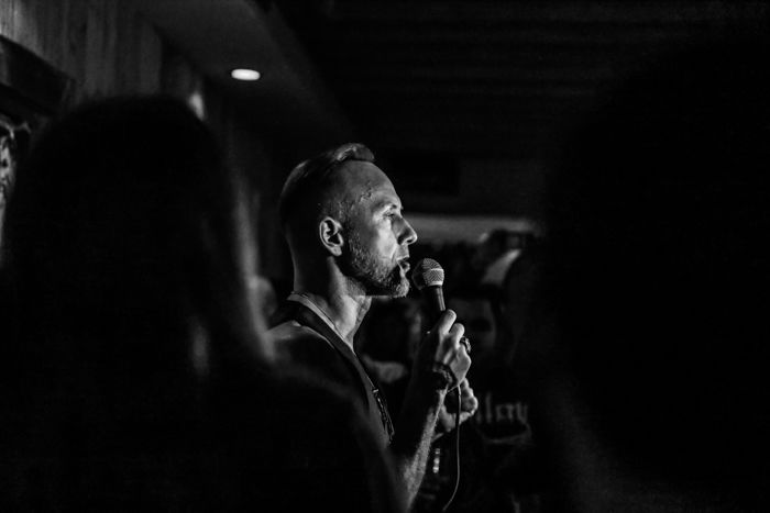 A black and white event photography shot of a man speaking or singing into a microphone onstage