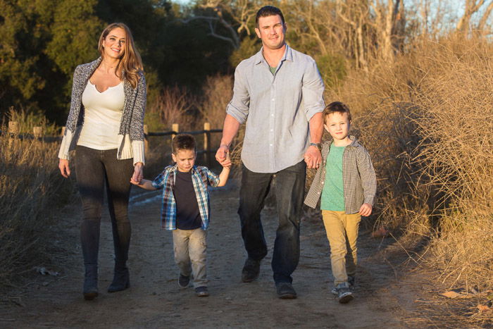 A portrait of a family of 4 smiling and holding hands while walking in the countryside