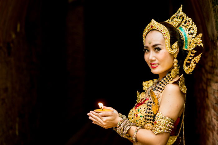 A beautifully costumed smiling thai dancer poses aginst a dark figure to ground background