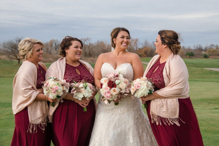 A bride posing with her bridesmaids in deep red gowns, holding bouquets, outdoors on a sunny day