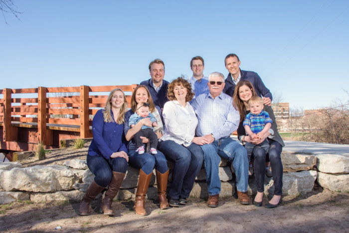 A family group photo outdoors on a bright day, family wearing jeans and boots, a wood bridge and blue sky behind them