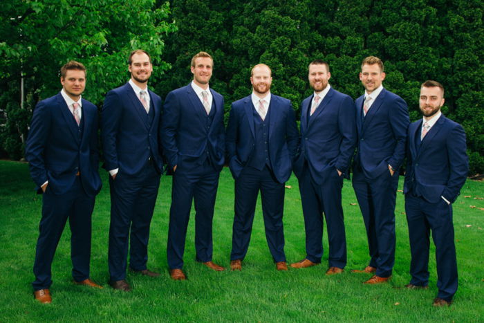 A group of groomsmen in blue black suits, in a curved line, posing in a grassy field, dark trees behind them