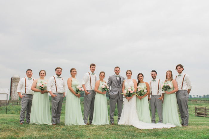 A wedding entourage posing in a grassy field, bridesmaids in mint gowns, groomsmen wearing suspenders