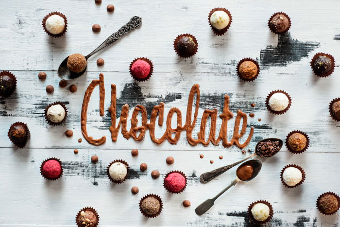 Word chocolate made from glazing with chocolates, candies and tea spoons on a white wooden background. Food typography concept. Sweet flatlay.