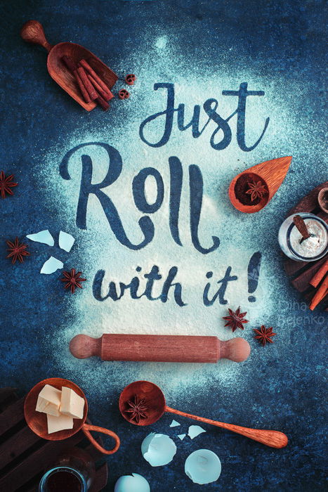 Just roll with it. Lettering in flour, surrounded by baking materials: rolling pin, wooden spoon, ingredients, on a dark blue teal background