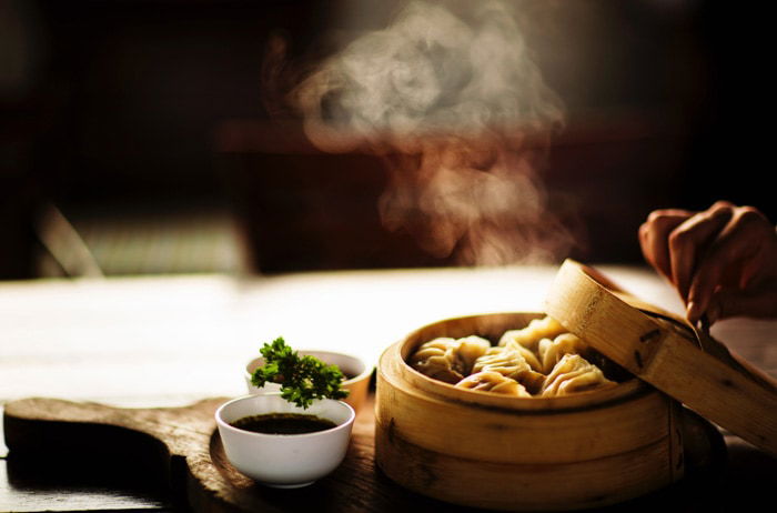 A person opening the lid on a steaming bowl of dumplings, beside two small bowls of sauce on a wooden chopping board 