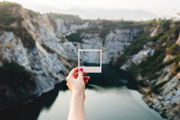 A photographer holding a Polaroid frame out against a mountainous landscape over a lake 