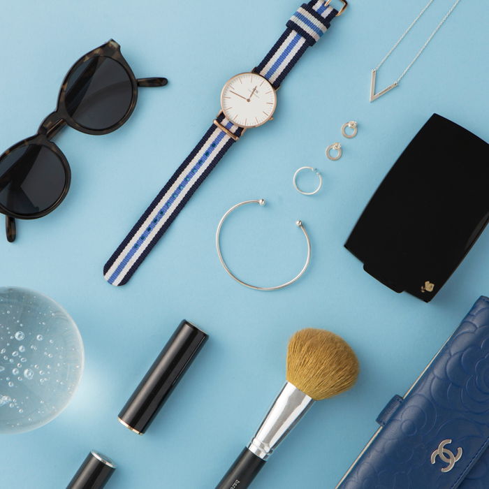 flatlay of woman's accessories on a light blue background: watch, earrings, sunglasses, brush, wallet