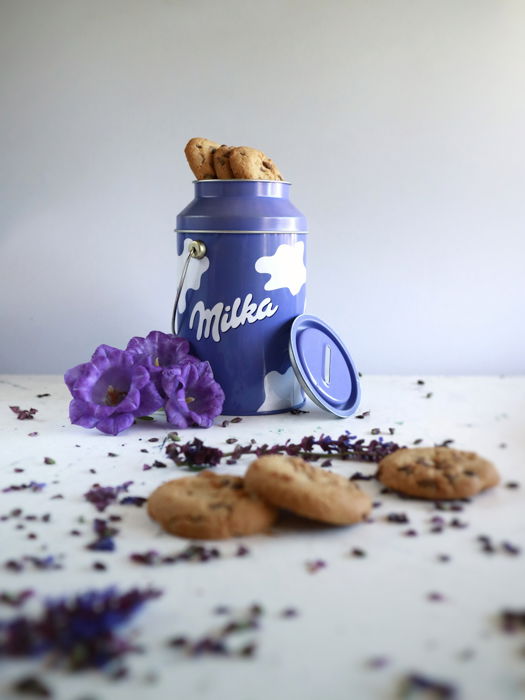 purple and white tin of cookies, purple flowers next to it, cookie and chocolate crumbs on the table