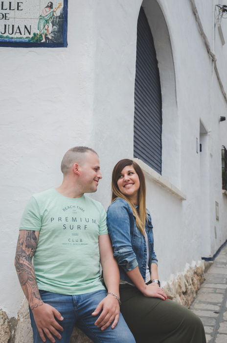 man and woman sitting at corner of a white building, smiling at each other lovingly