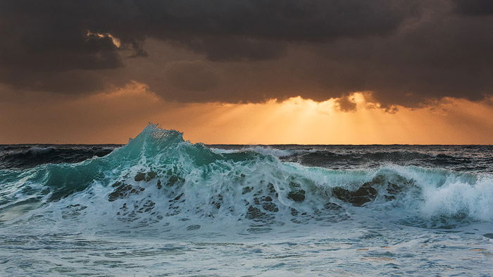 foamy waves crashing onto the shore, golden sun beams peeking from the dark clouds on the horizon. Shutter speed is 1/50 sec