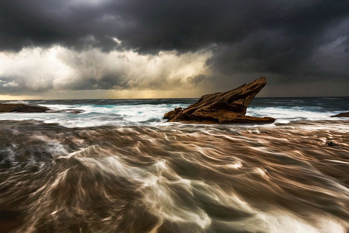 rock platform outcrop in the choppy sea waters, dark cloudy skies, a fading golden sunset on the horizon - water photography settings