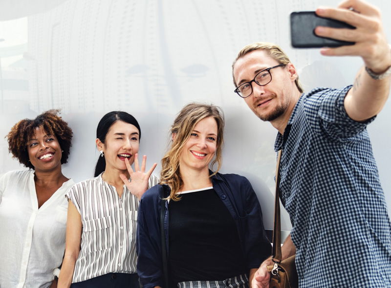 A man holding up a smartphone camera to take a selfie with three woman against a white wall with a faint reflection