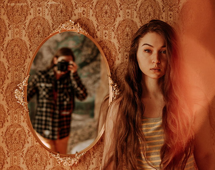 An artistic indoor portrait of a female model posing beside a mirror in which the photographer is reflected, examples of great profile pictures