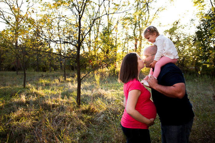 A pregnant woman and man with small child on his shoulders kisssing in a natural relaxed pose in a forest setting