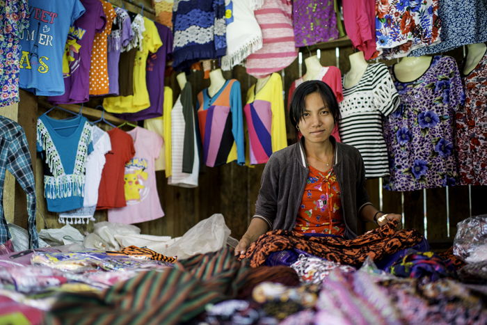 A female market vendor at her stall, with a busy cluttered background of colored material and clothes 