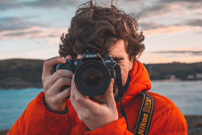 A man in orange jacket holding a Nikon DSLR camera 