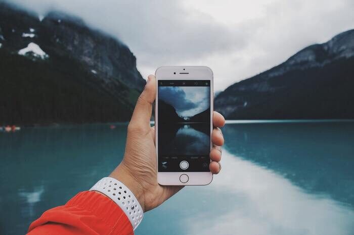 A hand holding up an iPhone with its camera app open showing a landscape scene of a mountain and lake