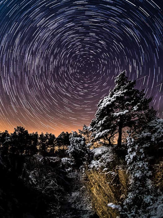 A lone pine tree on a cliff under a classic star trail centered on Polaris. 