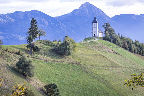 A small white church perched on the top of a grassy hill with a mountain in the background, surrounded by green fields and trees. 