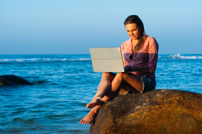 A girl using her laptop while sitting on a rock at the beach - bad stock photos of people