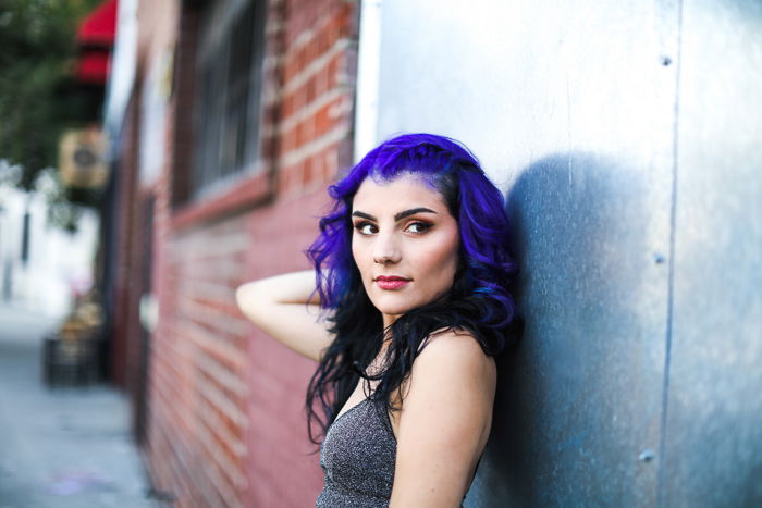 A portrait of a purple hair photography model posing by a brick wall