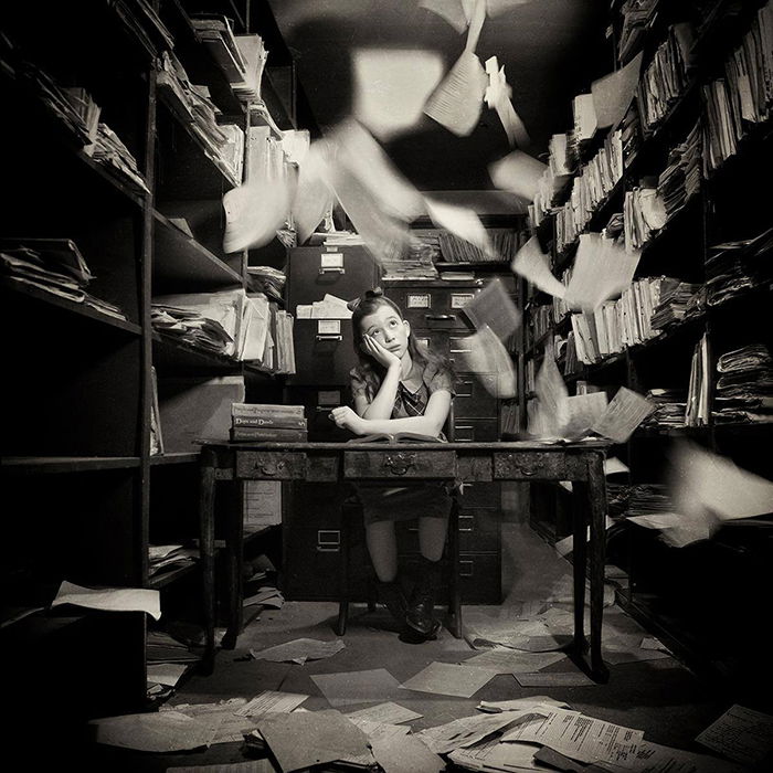 Conceptual black-and-white portrait of a young girl sitting in an office with papers flying by fine art photographer Carolyn Hampton