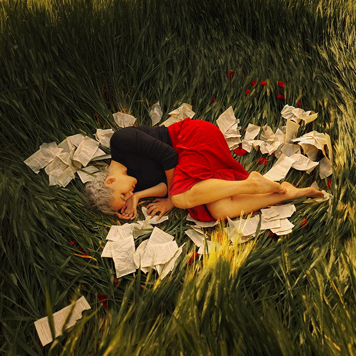 A female model curled up in grass surrounded by paper by fine art photographer Masha Sardari